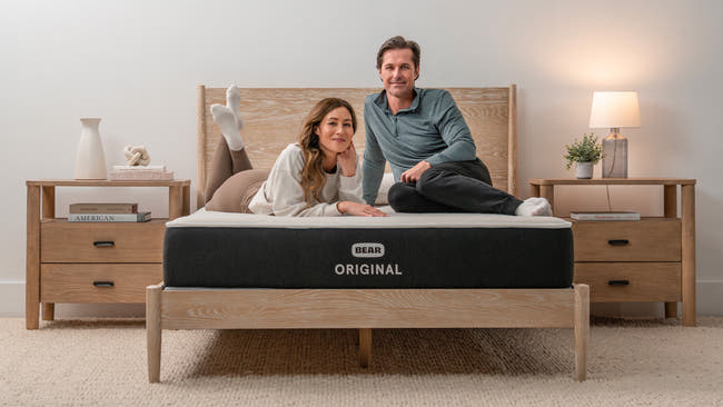 Couple sitting together on Bear Original mattress in warm bedroom with oak bed frame and nightstands for The Mattress Shop, showcasing product image Bear Original on the page: Best Memory Foam Mattresses page.
