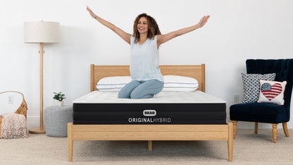 Smiling woman sitting on Bear Original Hybrid mattress in casual bedroom with bamboo bed frame and navy chair
