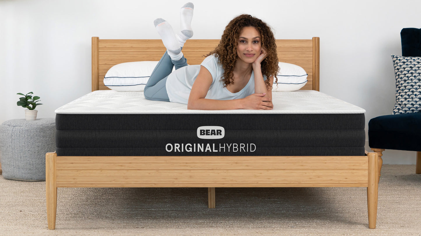 Woman lying on Bear Original Hybrid mattress smiling in bright bedroom with bamboo bed frame for The Mattress Shop, showcasing product image for Bear Original Hybrid on the product page.