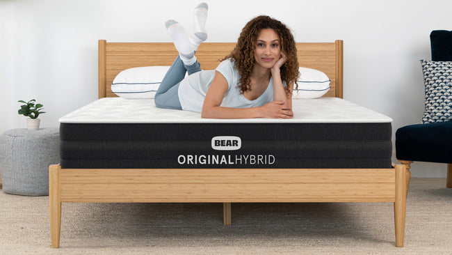 Woman lying on Bear Original Hybrid mattress smiling in bright bedroom with bamboo bed frame for The Mattress Shop, showcasing product thumbnail image for Bear Original Hybrid on the product page.