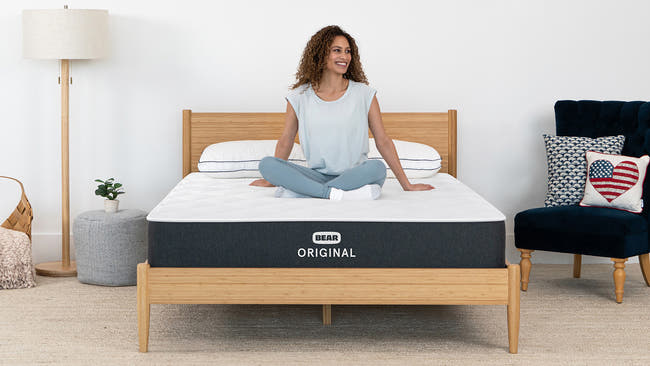 Woman sitting cross-legged on Bear Original mattress in bright bedroom with bamboo bed frame for The Mattress Shop, showcasing product image Bear Original on the page: Best Memory Foam Mattresses page.