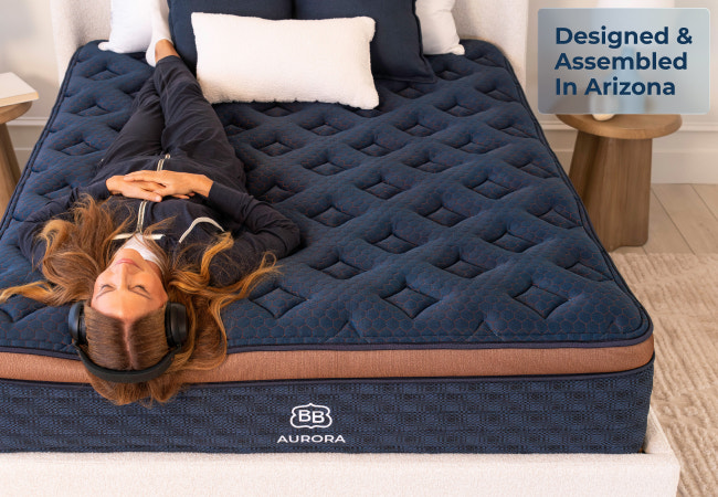 Brooklyn Bedding Aurora mattress, woman lying on navy blue quilted surface with headphones, designed and assembled in Arizona. for The Mattress Shop, showcasing product image Brooklyn Bedding Aurora Luxe Cooling on the page: Best Hybrid Mattresses page.