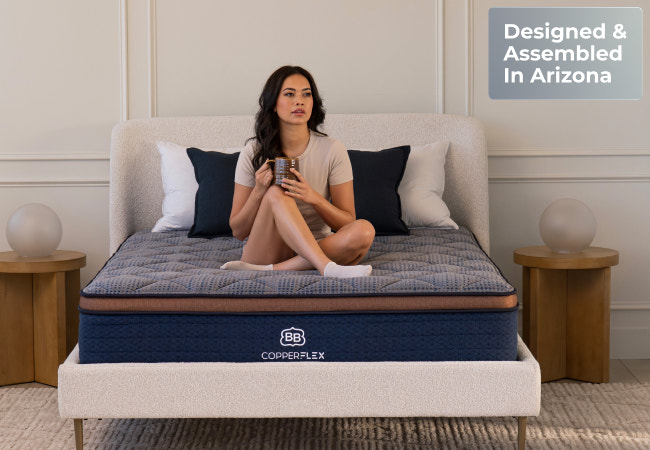 Brooklyn Bedding CopperFlex navy blue quilted pillow top mattress, woman sitting cross-legged on bed, designed and assembled in Arizona for The Mattress Shop, showcasing product image Brooklyn Bedding CopperFlex® on the page: Best Hybrid Mattresses - Quiz Prominent CTA page.