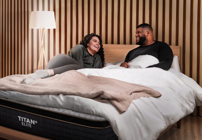 Brooklyn Bedding Titan Plus Elite mattress, couple laughing together in bed in modern wood panel bedroom for The Mattress Shop, showcasing product thumbnail image for Brooklyn Bedding Titan® Plus Elite on the product page.