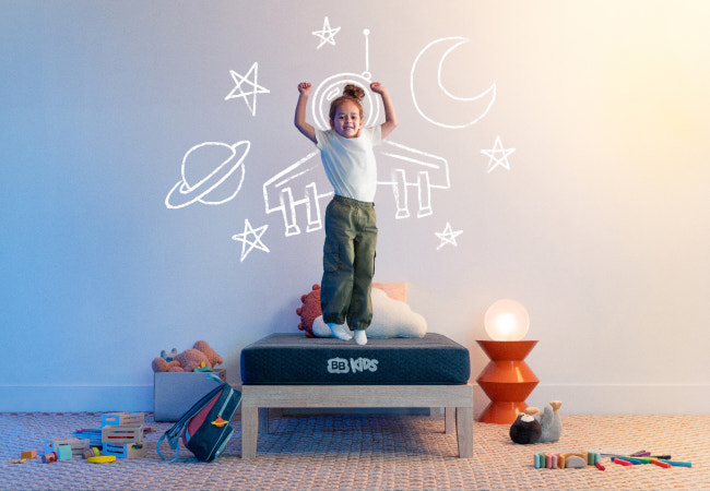 Brooklyn Bedding BB Kids mattress, child jumping on bed with whimsical space rocket drawing on wall behind her. for The Mattress Shop, showcasing product image Brooklyn Bedding BB Kids on the page: Best Mattresses for Kids page.