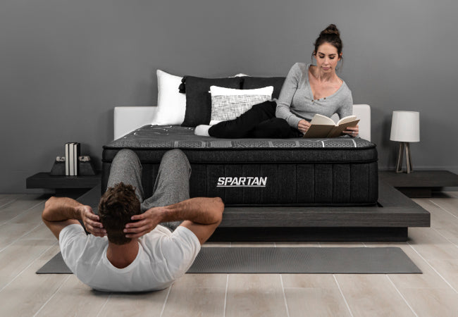 Brooklyn Bedding Spartan mattress, workout-inspired recovery sleep, man doing sit-ups while woman reads on bed. for The Mattress Shop, showcasing product image for Brooklyn Bedding Spartan on the product page.