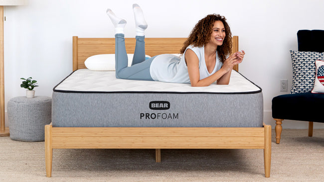 Smiling woman lying on Bear Pro Foam mattress in bright bedroom with bamboo bed frame and navy chair for The Mattress Shop, showcasing product thumbnail image for Bear Pro Foam on the product page.
