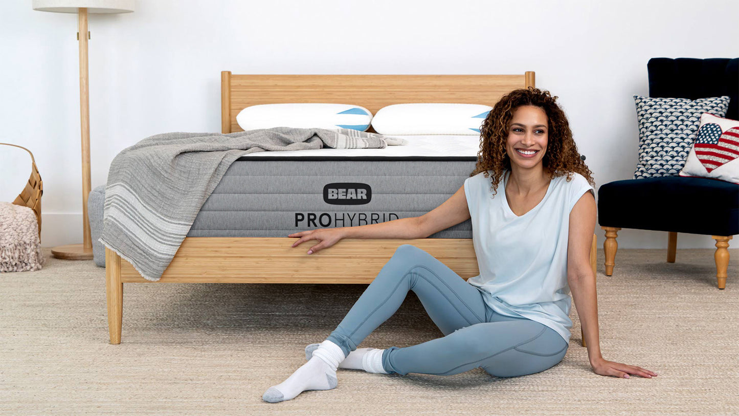 Woman in activewear sitting beside Bear Pro Hybrid Mattress on natural wood bed frame in bright bedroom for The Mattress Shop, showcasing product image for Bear Pro Hybrid on the product page.