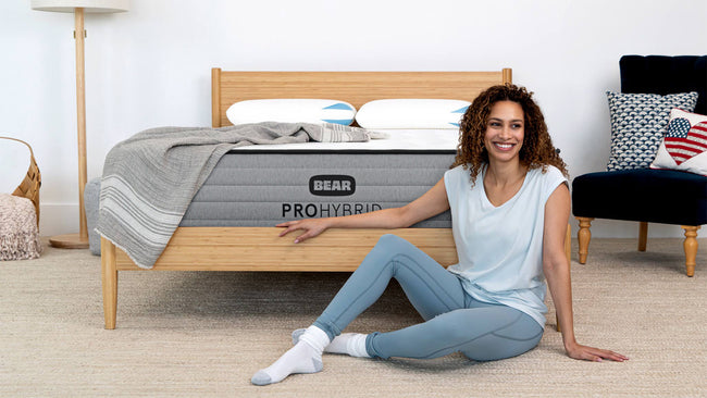 Woman in activewear sitting beside Bear Pro Hybrid Mattress on natural wood bed frame in bright bedroom for The Mattress Shop, showcasing product thumbnail image for Bear Pro Hybrid on the product page.